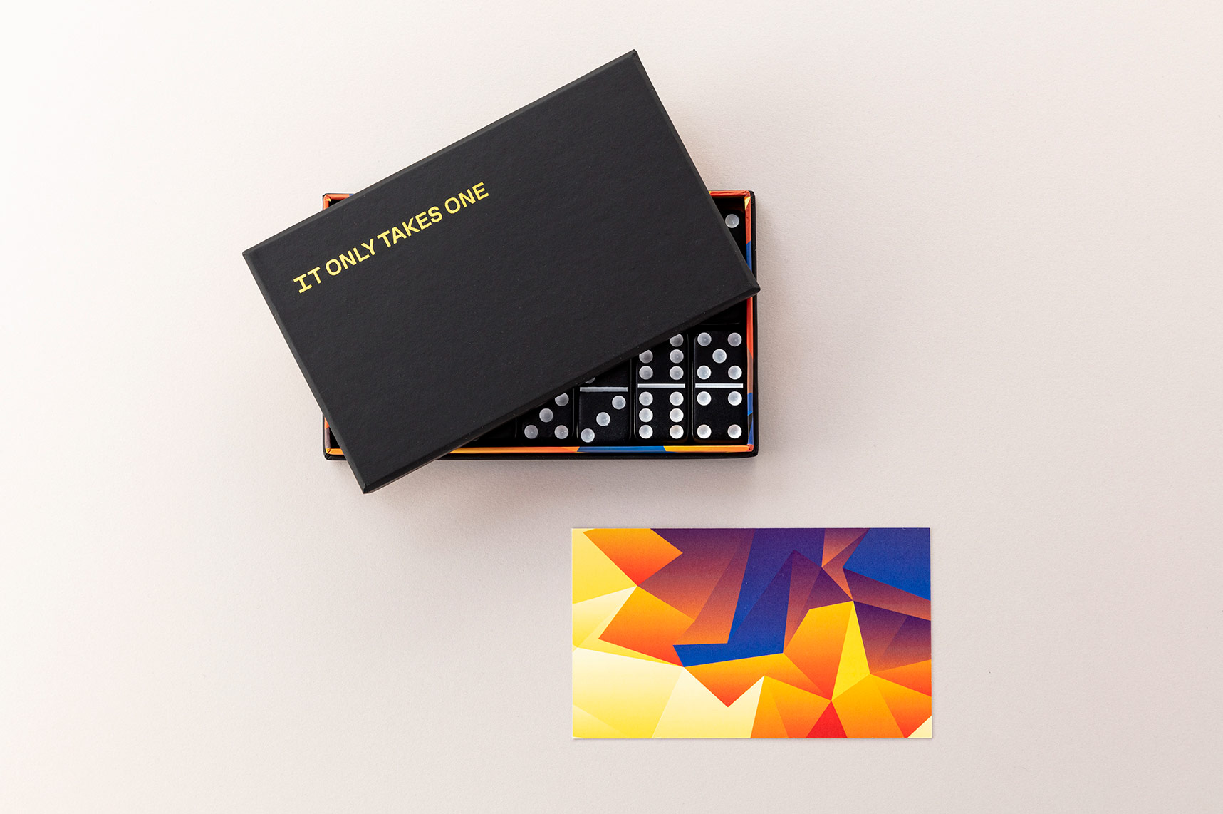 Black box with IT ONLY TAKES ONE printed on the top in gold foil and a set of dominoes are shone packaged inside.